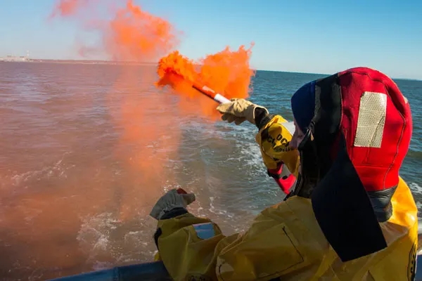 Woman at sea signalling for help with orange hand smoke flare while wearing an immersion suit